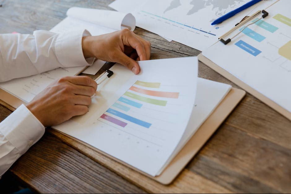 Person in a white shirt reviews printed bar charts, a "Brainstorming" flowchart, and a world map, planning AI chatbot development services on a wooden table.
