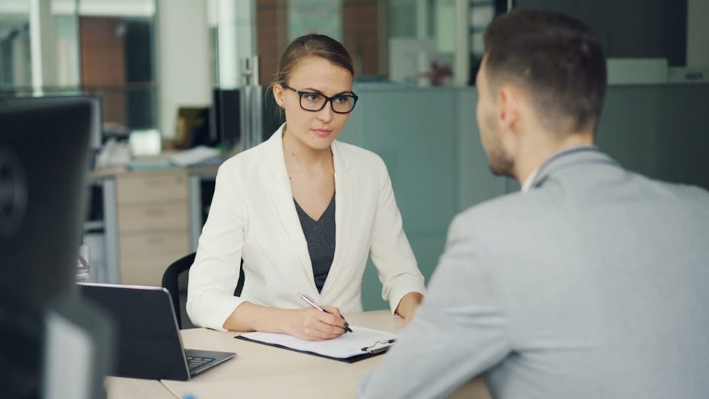 A professional interaction between a woman and a man in an office. The woman, wearing a white blazer and glasses, takes notes on a clipboard while engaging with the man, who is seen from behind. The setting includes a desk, a laptop, and a neutral-toned office background, depicting the creation of a B2B buyer persona.
