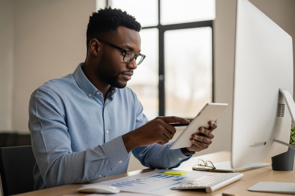 A Black man in a light blue shirt sits at a desk, focused on a tablet. The desk features papers with charts, a keyboard, a mouse, and a potted plant. A computer monitor is visible, and the background includes a window with natural light, representing a B2B buyer persona in action.