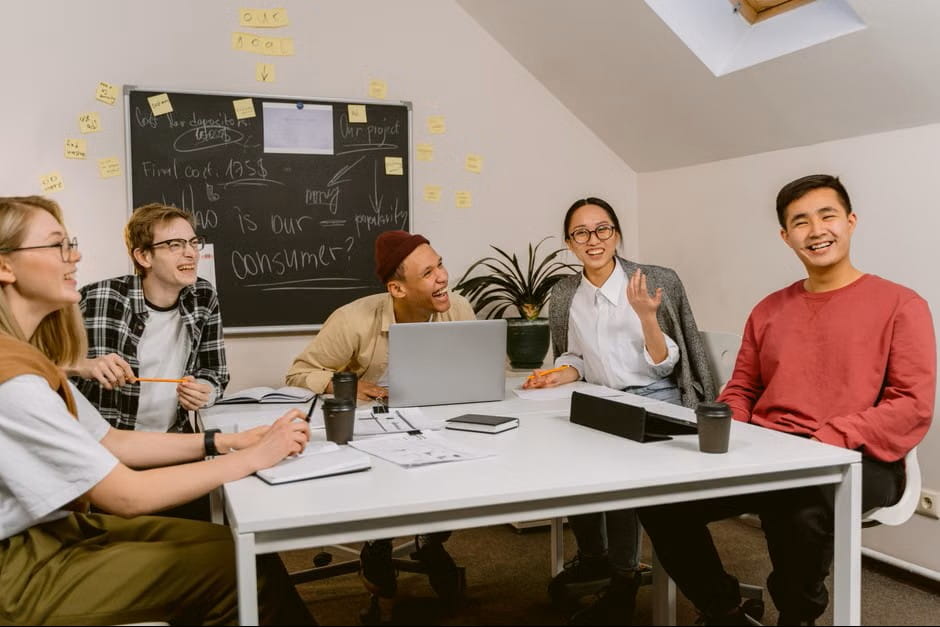 Five young professionals laugh and brainstorm employee engagement ideas around a conference table filled with laptops, coffee cups, and a chalkboard in a bright modern office.