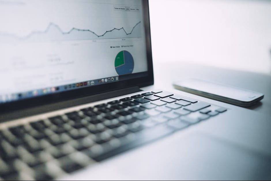Laptop showing a web analytics dashboard with a line graph and pie chart comparing new and returning visitors, next to a smartphone on a clean white desk.