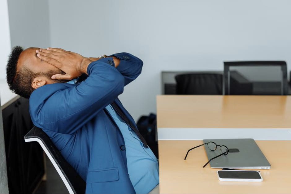 A man in a blue suit leans back in his office chair with his hands over his face, showing the stress and burnout that better employee engagement ideas can help prevent.