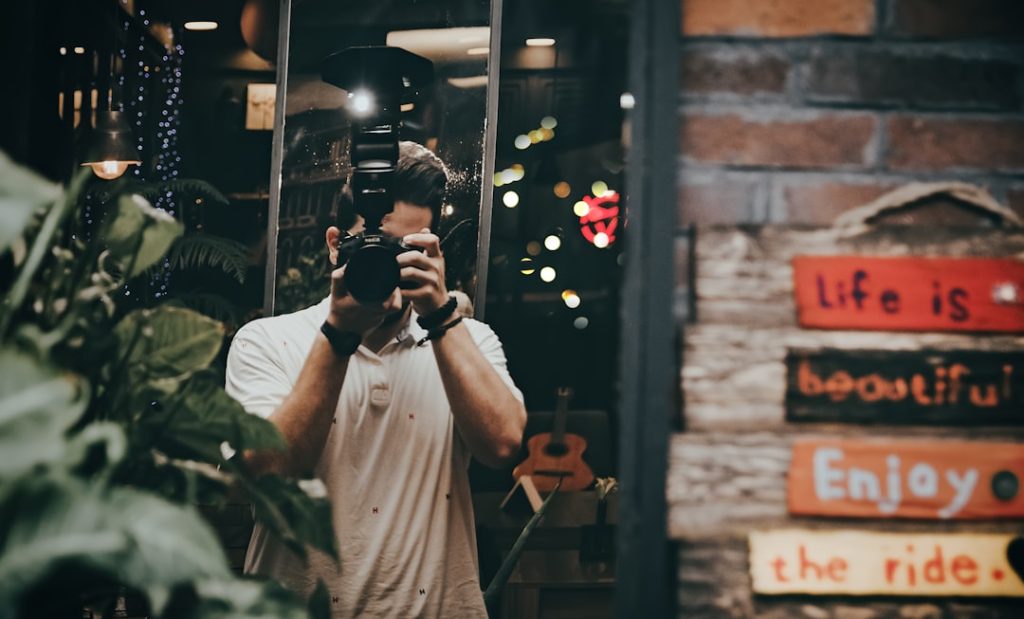 A mirror selfie of a person holding a DSLR camera with a flash, taken in a dimly lit space while learning how to use social media for small business. The background features bokeh lights, plants, and a guitar. A brick wall with colorful plaques reading 'Life is beautiful, enjoy the ride' is visible on the right.