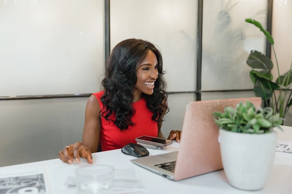 A smiling Black woman with long, dark curly hair sits at a white desk in a modern office, learning how to use social media for small business on her laptop with a pink protective cover. The desk features a black mouse, a smartphone on a wireless charging pad, and potted plants. The background includes frosted glass panels and a larger green plant.