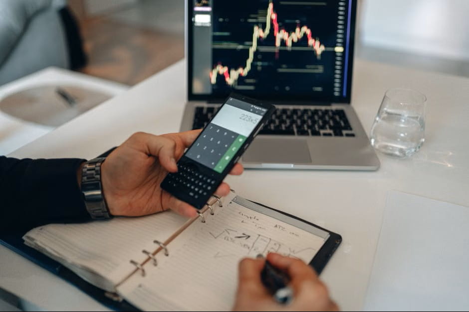 Person using a phone calculator and laptop stock chart for profit margin calculator planning and notes.