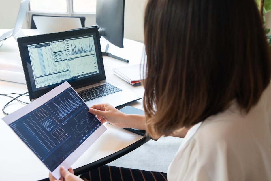 Woman analyzing Small Business Administration data on a laptop and printed sheet in a modern office.