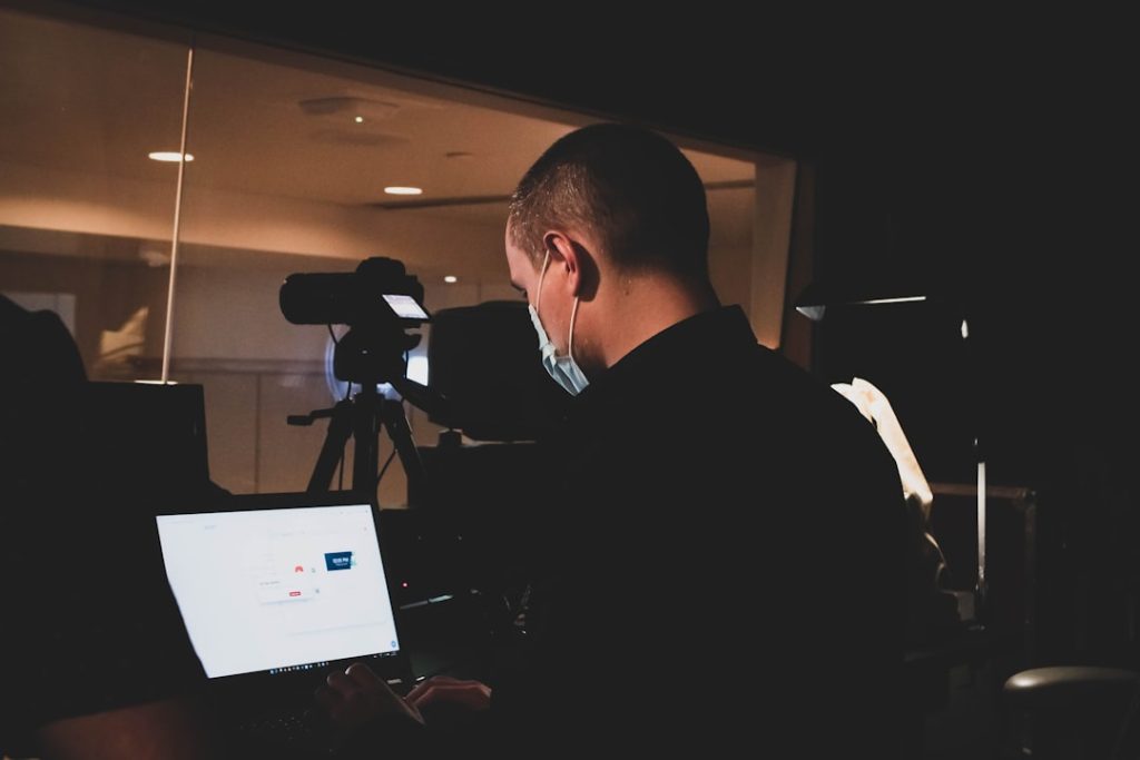 A person wearing a face mask sits at a desk, typing on a laptop while applying some helpful video conference tips for their next meeting. A professional video camera on a tripod is positioned behind them. The dimly lit background includes a glass partition reflecting ceiling lights.