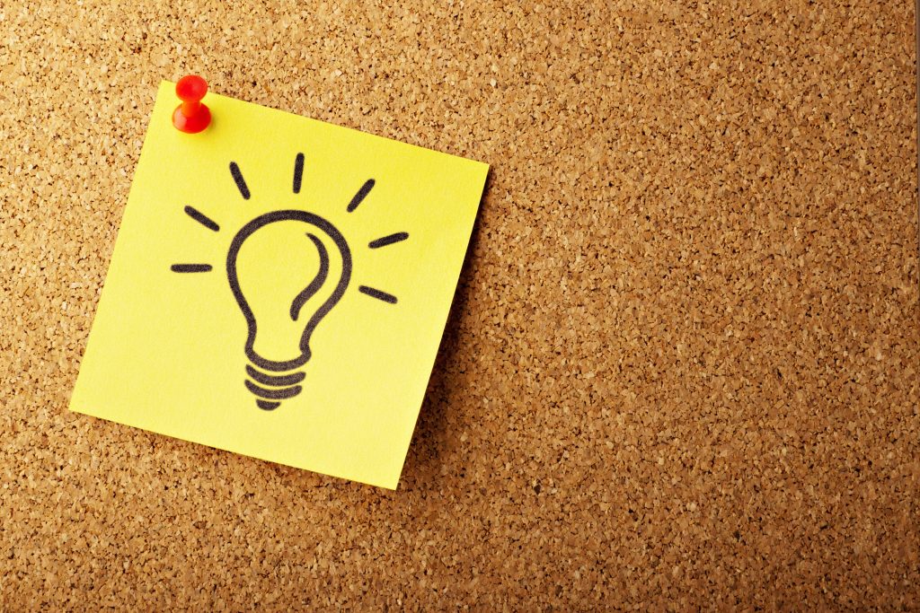 Close-up of a bright yellow sticky note pinned to a textured cork board with a red thumbtack. The sticky note features a simple black outline drawing of a lightbulb with radiating lines, symbolizing an idea or inspiration for networking events Philadelphia. The cork board provides a warm, neutral background, emphasizing the vibrant colors of the note and thumbtack.