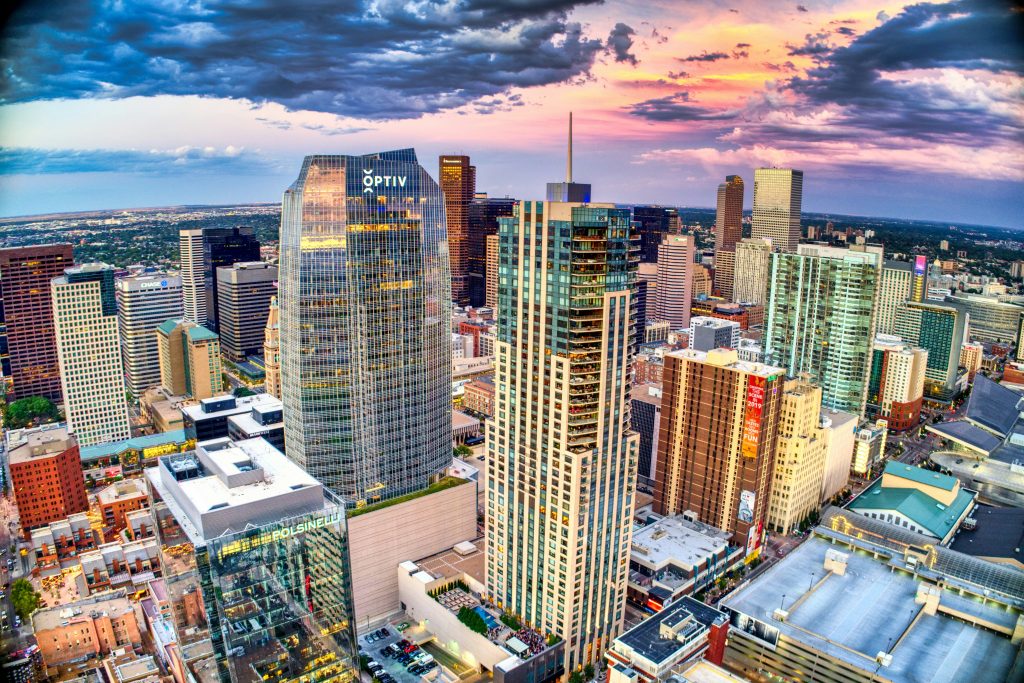 An aerial view of Denver's city skyline at sunrise or sunset, featuring a dramatic sky with hues of blue, pink, and orange. The cityscape includes a mix of modern skyscrapers and historic buildings, with prominent structures like the glass 'OPTIV' building, the 'CHASE' building with horizontal stripes, and a brown high-rise displaying a banner for 'CELEBRATING THE SCENE 2019.' The foreground shows rooftops with green spaces and amenities, while the background reveals a golden-domed building and a flat horizon. The illuminated windows and reflective glass facades add vibrancy to the bustling urban scene, a perfect setting for networking events Denver.
