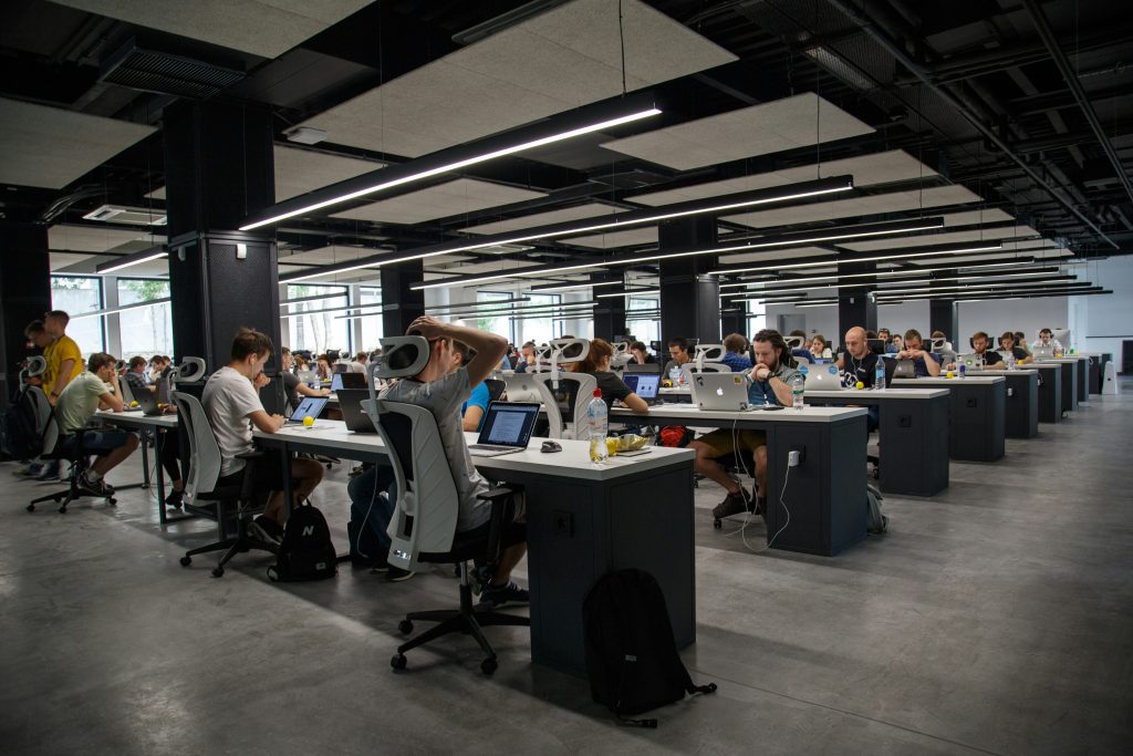 A modern open-plan office with rows of white desks, ergonomic chairs, and people working on laptops, illuminated by LED ceiling lights, with a casual and collaborative atmosphere, where discussions about exit interview questions occasionally take place.