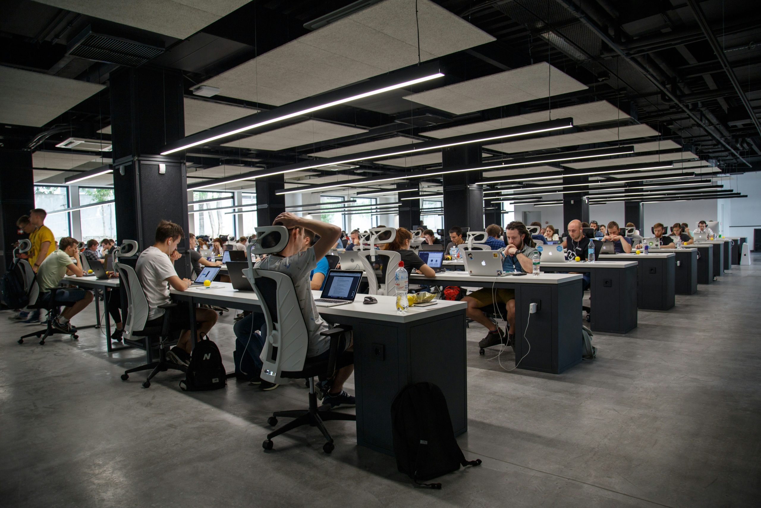 A modern open-plan office with rows of white desks, ergonomic chairs, and people working on laptops, illuminated by LED ceiling lights, with a casual and collaborative atmosphere, where discussions about exit interview questions occasionally take place.