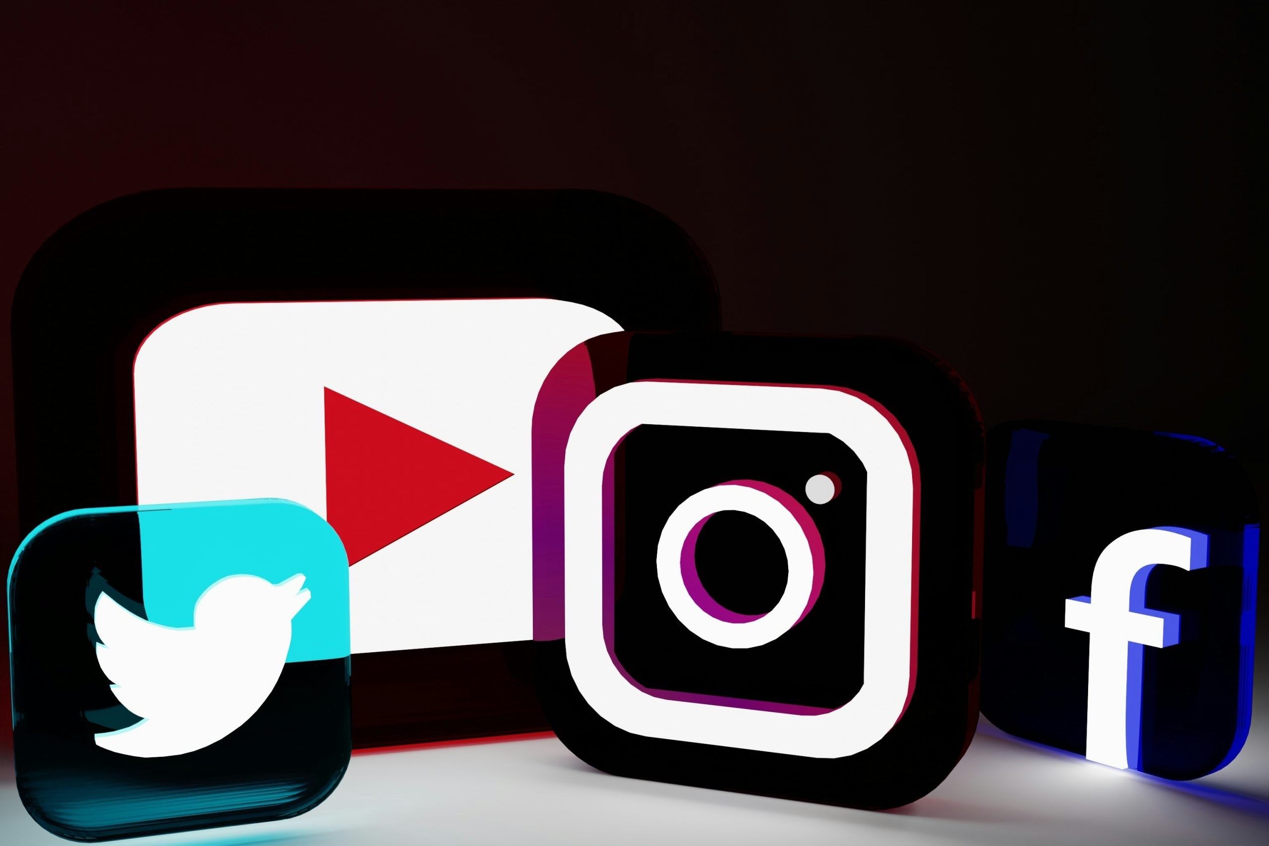Four glowing 3D social media logos arranged diagonally on a dark background. The Twitter logo, a white bird on a turquoise square, glows blue in the bottom left. The YouTube logo, a white play button on a red rectangle, glows red and purple. The Instagram logo, a white camera icon, glows purple-pink. The Facebook logo, a white 'f', glows blue on the far right. The dark background enhances the vibrant, illuminated logos, creating a modern and digital aesthetic. This visual represents the best way to keep up on social media trends.