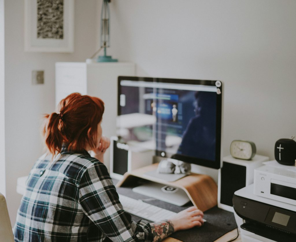 A person with reddish-brown hair in a ponytail, wearing a blue and white plaid shirt, sits at a desk researching the things needed in a business website on a computer. The desk features a large monitor on a wooden stand, a white keyboard, a dark gray mousepad, and a computer mouse. Surrounding the workspace are items like speakers, a vintage-style green alarm clock, a white printer, and a black portable speaker. The background includes a light-colored wall, a framed picture, a turquoise desk lamp, and a white shelf. The scene is softly lit with natural light, creating a calm and focused atmosphere.