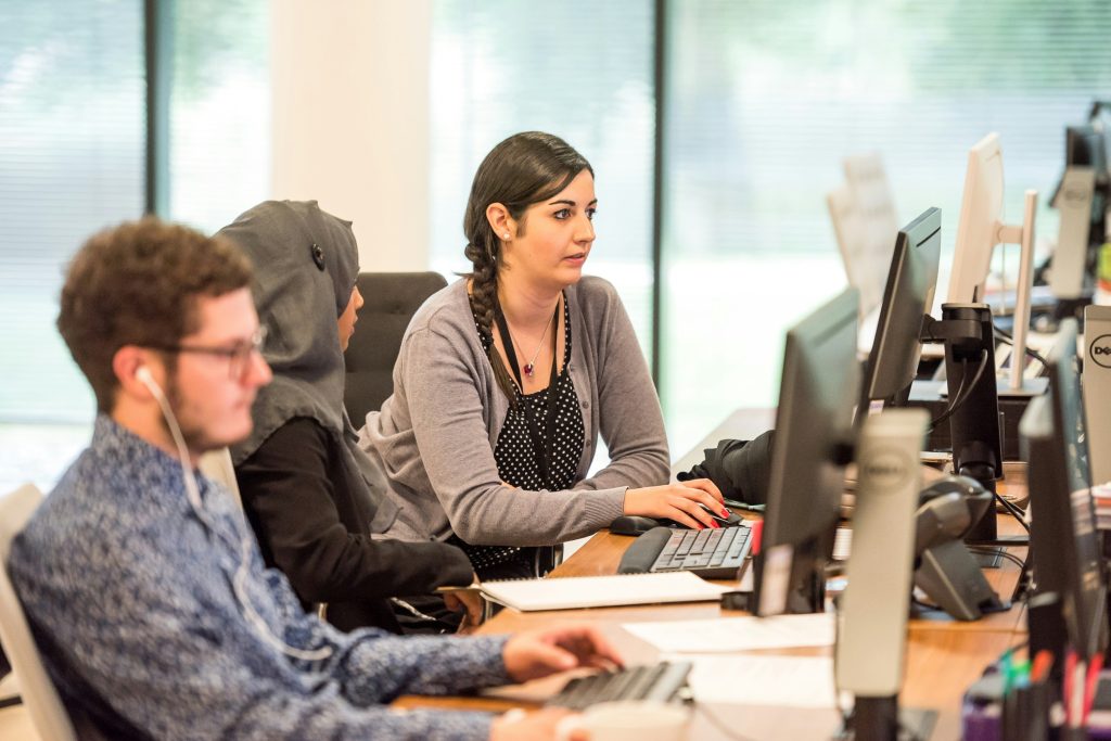Three people seated at a shared wooden desk, working on computer monitors in a bright office space with large windows and natural light, discussing exit interview questions.