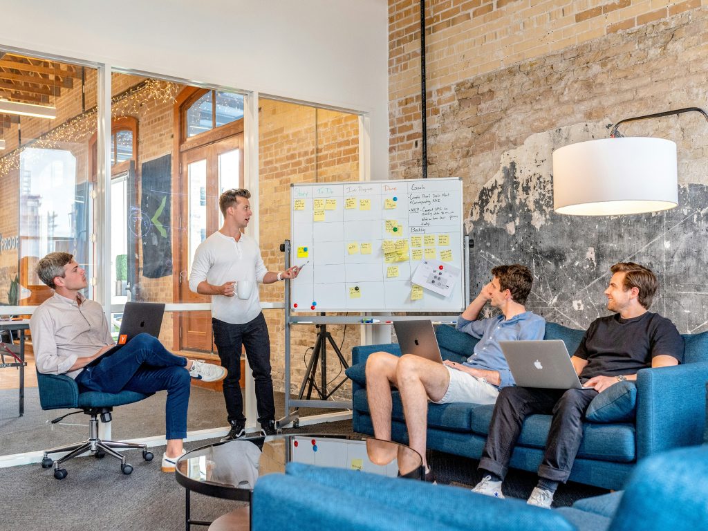 Four men in a modern office space engaged in a team meeting. One man stands in front of a whiteboard divided into columns labeled 'Story,' 'To Do,' 'In Progress,' 'Done,' 'Goals,' and 'Backlog,' covered with sticky notes and handwritten text. He gestures with a pen while holding a white mug. Two men sit on a blue sofa with laptops, and another sits on an office chair with a laptop. The setting features exposed brick walls, glass partitions, string lights, and a round glass coffee table, creating a collaborative and creative atmosphere perfect for cheap small business marketing agencies.