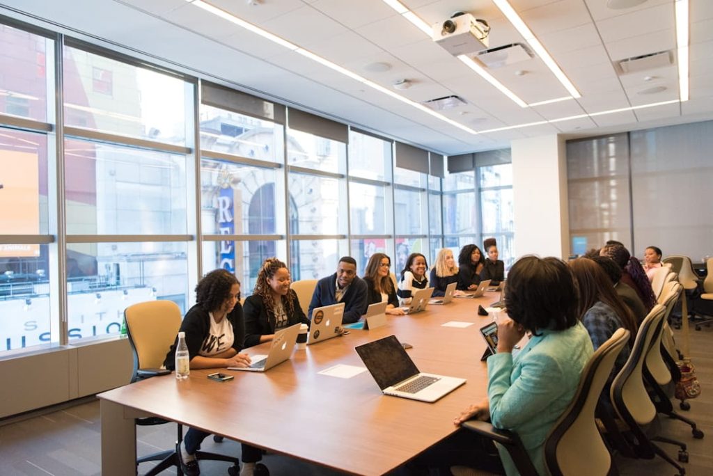 A diverse group of professionals in a modern conference room, seated around a large table with laptops, water bottles, and coffee cups. The room has floor-to-ceiling windows offering a view of city buildings, and a ceiling-mounted projector suggests a collaborative meeting environment for B2B lead scoring.