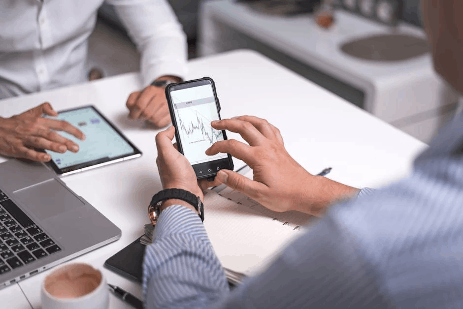 A collaborative office scene featuring two professionals analyzing financial data for a b2b retargeting campaign on multiple devices, including a smartphone displaying a volatile market chart, a tablet, and a laptop. The setting includes a white desk with a coffee cup, pen, and a blurred office background.