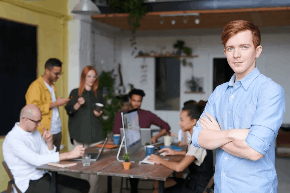 A modern, open-plan office with a diverse group of professionals working and conversing. The foreground features a confident young man with red hair and a blue shirt, standing with arms crossed. The background includes a communal table with laptops, coffee mugs, and team members collaborating on a b2b retargeting campaign.