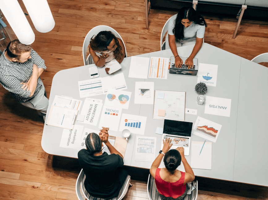 A high-angle view of a collaborative marketing meeting around a pentagonal table. Team members are engaged with laptops, documents, and charts labeled "Marketing Strategy," "Trends," and "Analysis" for a b2b retargeting campaign. The setting includes pendant lights, white chairs, and a mix of physical and digital tools.