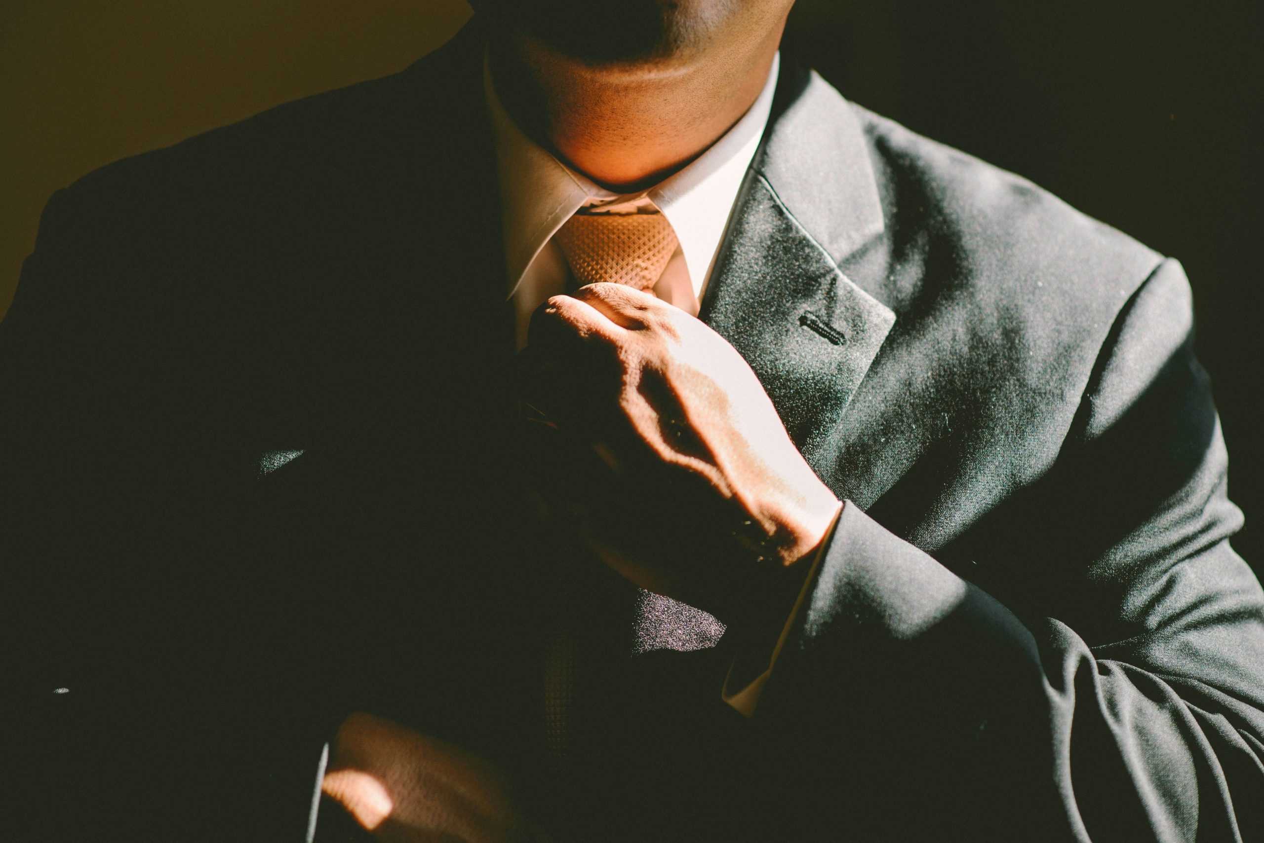 A close-up, medium shot of a small business lawyer adjusting their tie. The individual is dressed in formal attire, featuring a dark suit jacket, a white shirt, and a patterned tie in a light gold or brown shade. The right hand is positioned at the tie knot, illuminated by warm, dramatic lighting that highlights the textures of the fabric and skin. The background is completely dark, creating a sophisticated and formal atmosphere.