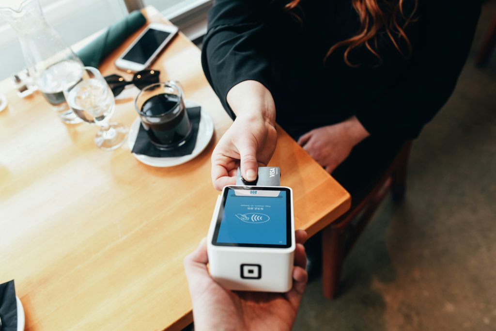 A customer hands a Visa credit card to another person holding a small white card reader displaying a contactless payment symbol and a charge of $23.85. The transaction takes place on a wooden table in a café setting, with a cup of coffee, a glass of water, a smartphone, and sunglasses visible on the table. The scene highlights a modern, convenient payment experience, showcasing the best credit card processing for small business.