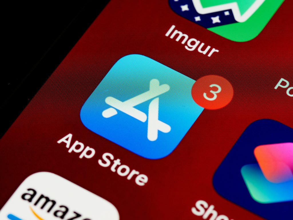 A close-up view of a smartphone screen displaying app icons on a dark red background. The App Store icon, a bright blue square with a white stylized 'A,' is prominently featured with a red notification badge showing the number '3.' Other partially visible icons include Imgur, Amazon, and possibly small business apps, arranged in a grid pattern. The screen exhibits a pixelated texture, characteristic of a magnified digital display.