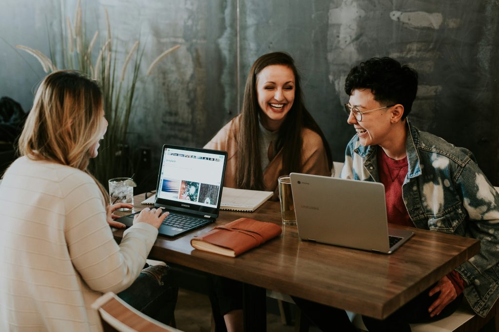 Three young adults sit around a wooden table, laughing and engaging in conversation, perhaps discussing their roles within an employee benefits corporation. On the left, a woman with long blonde hair types on a black Samsung laptop, with a clear drink and lime wedge beside her. In the middle, a woman with long brown hair laughs heartily, with an open spiral notebook in front of her. On the right, a person with short curly hair and gold-rimmed glasses laughs while seated in front of a silver Samsung laptop, with a glass mug of yellow liquid nearby. The background features a textured wall and ornamental grasses, creating a relaxed and collaborative atmosphere.