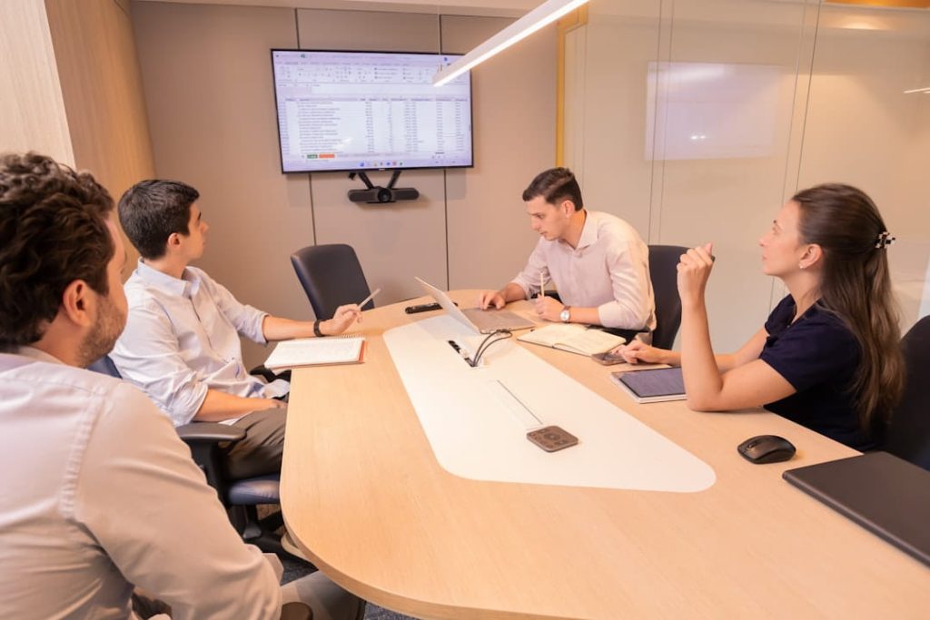 A modern conference room with four professionals seated around a light wooden table, engaged in discussion at one of many business analyst networking events. A large wall-mounted monitor displays a spreadsheet, suggesting a data-driven meeting. Laptops, notebooks, and other office supplies are scattered on the table.