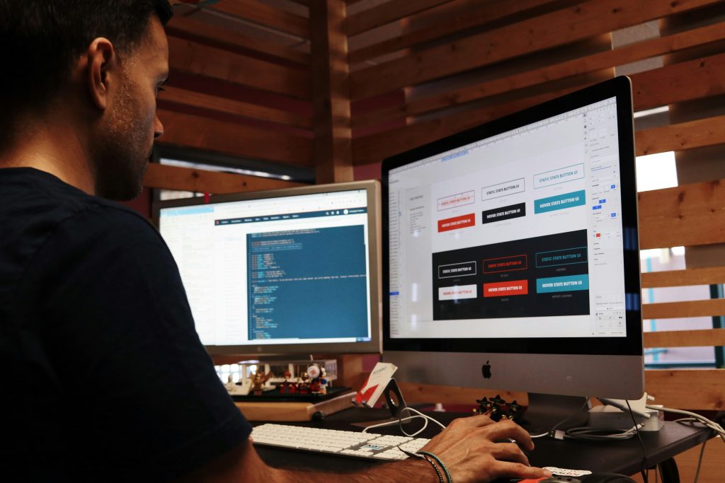 A male designer or developer working on a small business website design at a desk with two monitors. The larger monitor on the right displays UI design software with various button styles labeled 'STATIC STATE BUTTON UI' and 'HOVER STATE BUTTON UI,' showcasing different color schemes like white, black, red, and teal. The smaller monitor on the left shows lines of code on a dark background. The desk features a white keyboard, a black mouse, small figurines, and a card labeled 'ALVARO.' The background is a wooden slat wall with light peeking through. The man, wearing a dark t-shirt and bracelets, is focused on his work, with his right hand on the mouse.