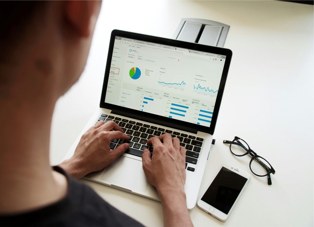 A workspace with a person at a desk, working on a laptop showing a dashboard interface. The screen displays data visualizations like a pie chart, line graphs, and bar charts. The left sidebar highlights 'Dashboard' alongside options like 'Home,' 'Reports,' and 'Settings.' On the desk, a smartphone and glasses sit next to the laptop. The setting reflects a professional environment focused on what spreadsheets to keep for a small business, data analysis, or reporting.
