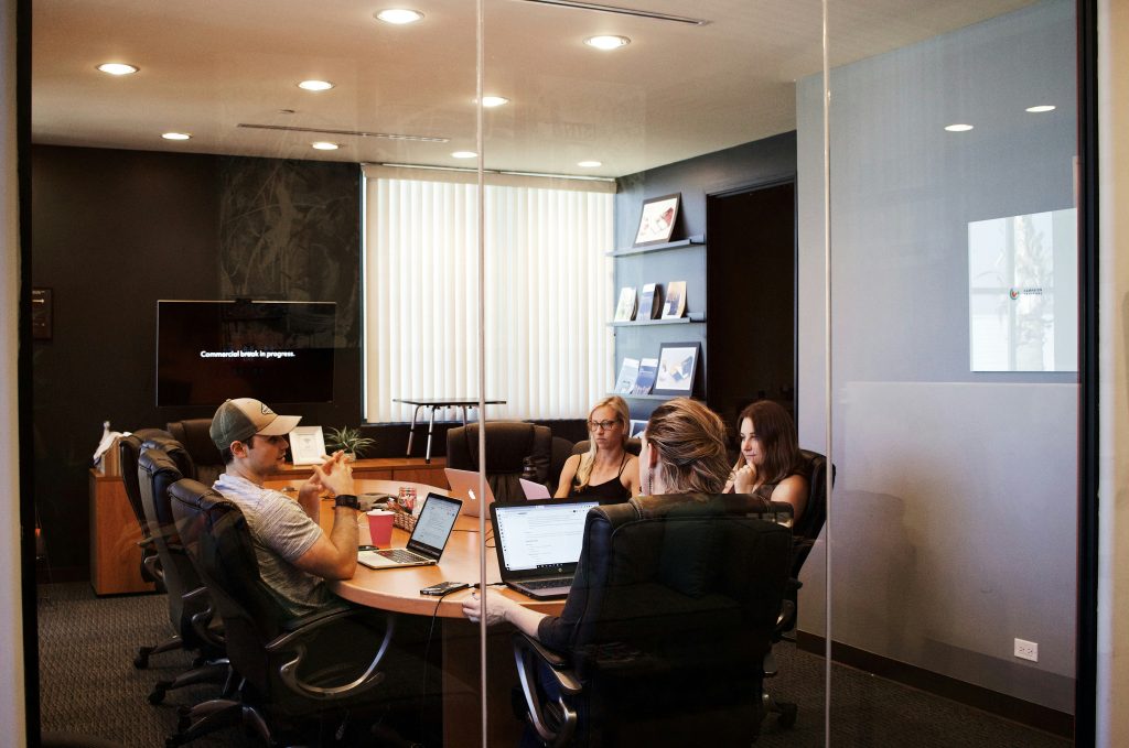A modern office meeting room viewed through a glass partition. Four individuals, including a small business lawyer, are seated around a large, round wooden table, each engaged with laptops. A man wearing a baseball cap and a smartwatch sits on the left, with a red cup, a woven basket, and a laptop in front of him. Two women are visible further around the table, one with light brown hair tied up and another with blonde hair and glasses, both focused on their laptops. The room features a dark wall with a mounted TV displaying 'Commercial break in progress,' vertical blinds covering a window, and a shelf unit holding books and framed pictures.