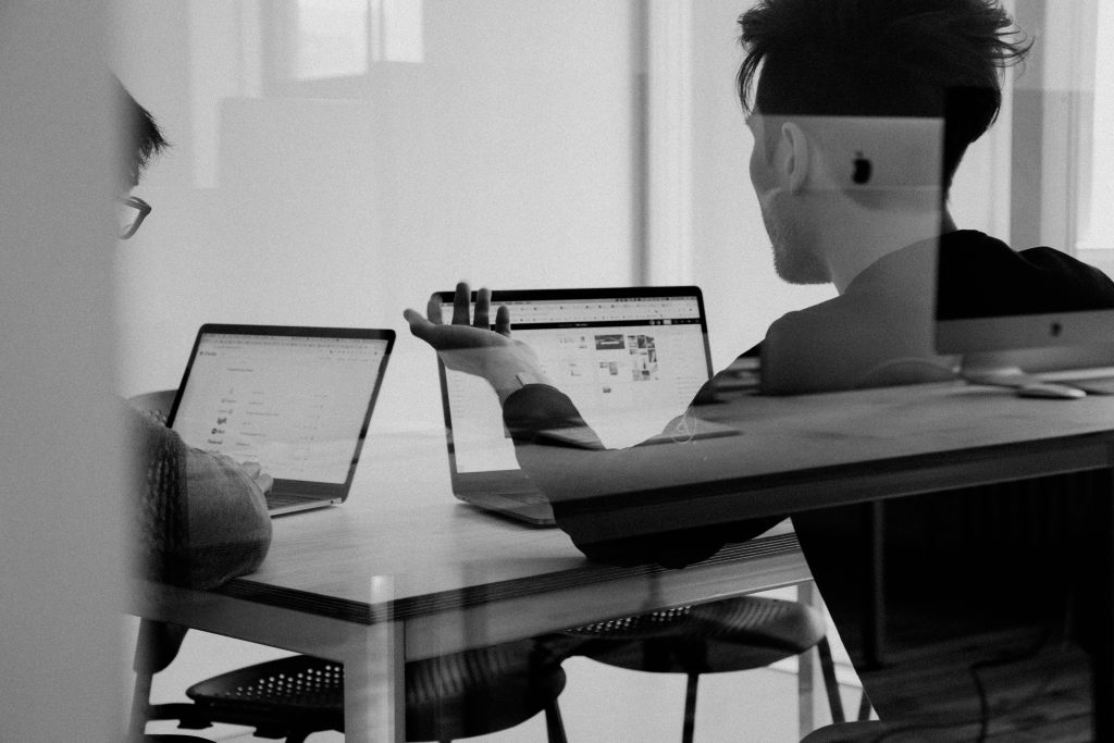 A black-and-white image shows two individuals working on laptops at a light-colored wooden table, viewed through a reflective surface that creates a double exposure effect. On the left, a person with dark hair and glasses focuses on their laptop, while on the right, a person with short hair and a beard gestures with one hand, as if explaining something for an AI Agency. The reflection overlays an Apple monitor logo and faint architectural details, adding depth and an artistic quality. The scene suggests collaboration in a modern, creative workspace.