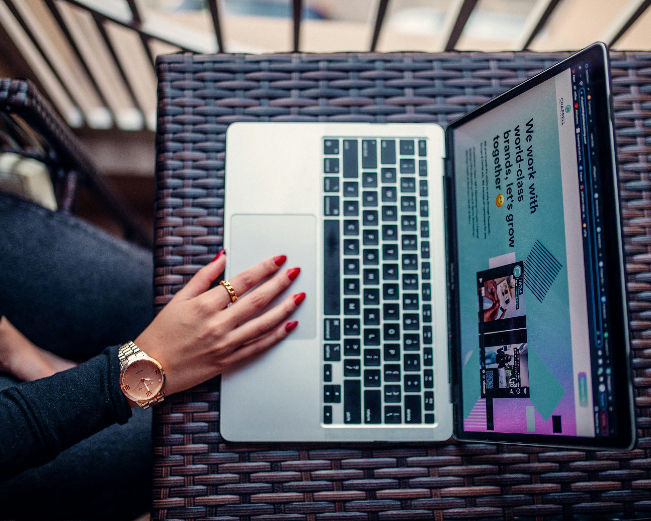 A stylish hand with red-painted nails, wearing a gold watch and rings, rests on a laptop trackpad on a dark wicker table, with the laptop screen displaying a webpage about brand collaboration and the best website builder for small business.