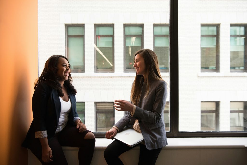 Two women sit side-by-side on a white windowsill, smiling and engaged in conversation. The woman on the left, with dark wavy hair, wears a dark blazer over a white top and rests her hand on her knee. The woman on the right, with long brown hair, wears a gray blazer over a light top and holds a silver laptop in her lap, gesturing with her other hand. Behind them, a large window reveals a white brick building with dark-framed windows. The scene suggests a bright, modern workspace, likely a spot for networking events in DC, and conveys a cheerful, collaborative atmosphere.