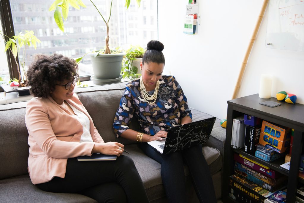 Two women seated on a gray sofa in a bright office setting with a large window overlooking a cityscape. The woman on the left, a small business consultant with curly hair and glasses, wears a pink blazer and holds a notebook, smiling as she looks to the right. The woman on the right, with a high bun and a cat-patterned blouse, types on a laptop with a marble-patterned cover. Behind them, potted plants line the windowsill, and a shelf filled with board games like 'Cards Against Humanity' and 'Pandemic' is visible. The space exudes a casual and creative atmosphere.