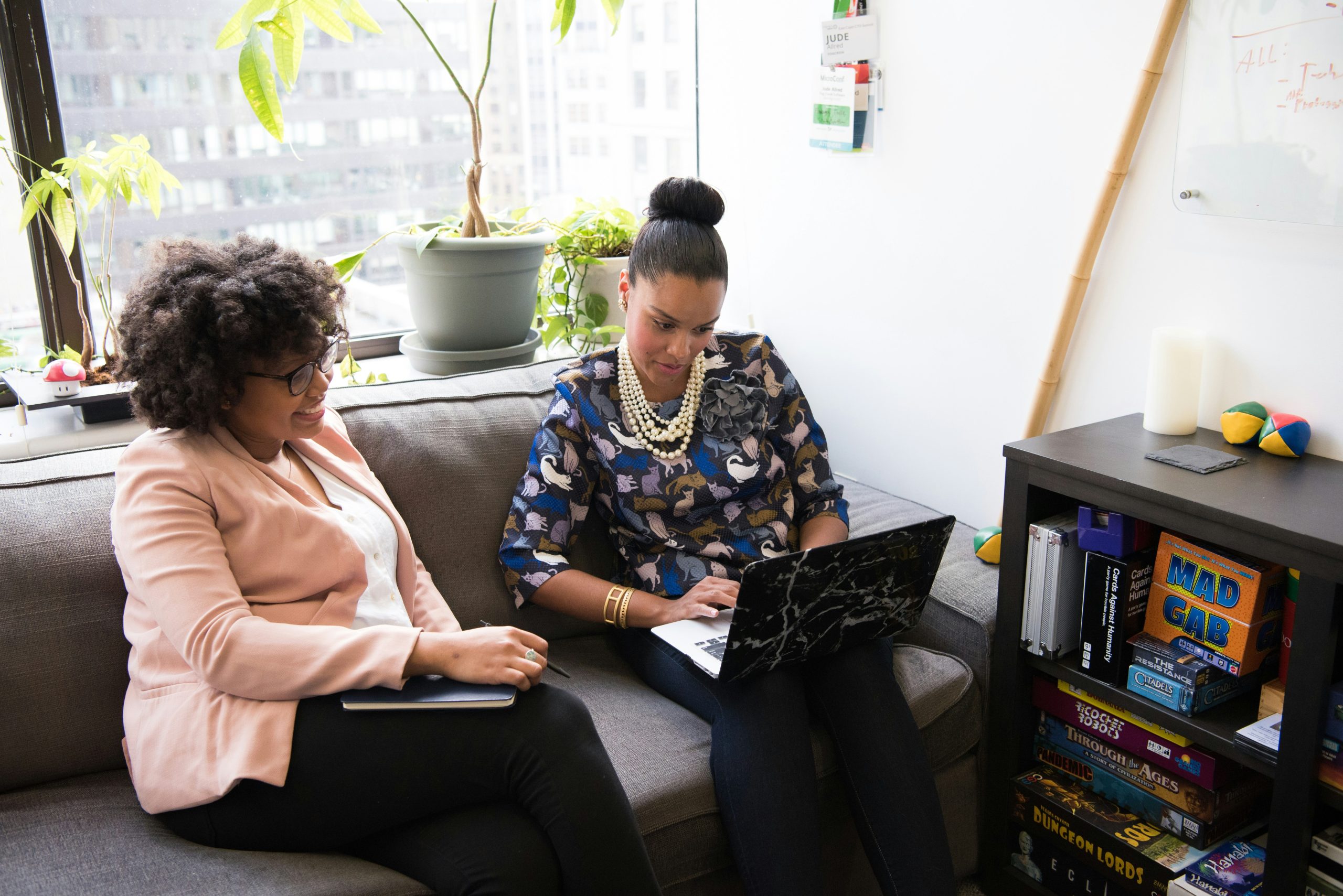 Two women seated on a gray sofa in a bright office setting with a large window overlooking a cityscape. The woman on the left, a small business consultant with curly hair and glasses, wears a pink blazer and holds a notebook, smiling as she looks to the right. The woman on the right, with a high bun and a cat-patterned blouse, types on a laptop with a marble-patterned cover. Behind them, potted plants line the windowsill, and a shelf filled with board games like 'Cards Against Humanity' and 'Pandemic' is visible. The space exudes a casual and creative atmosphere.