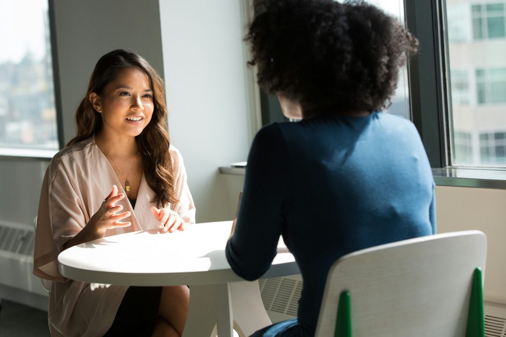 Two individuals seated at a white round table in a bright, modern office setting with large windows revealing a cityscape. The woman on the left, a small business consultant with medium brown wavy hair, smiles warmly and gestures with her left hand while wearing a light-colored top and a gold necklace. The person on the right, seen from behind, has dark curly hair and wears a dark blue shirt. Natural light streams in from the windows, creating a welcoming and professional atmosphere.