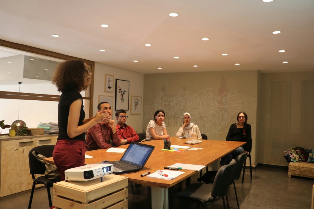 A modern meeting room with seven individuals engaged in a presentation. A woman with curly brown hair and glasses stands on the left, gesturing as she presents to an employee benefits corporation. Six attendees sit around a large wooden table, listening attentively. The table holds a laptop, colorful sticky notes, pens, a glass pitcher, and notebooks. A white Epson projector sits on a crate in front of the table. The background features a city map mural with one building highlighted in orange, framed artwork, and a cabinet with plants and a basket. The room is brightly lit with recessed ceiling lights, creating a professional and collaborative atmosphere.