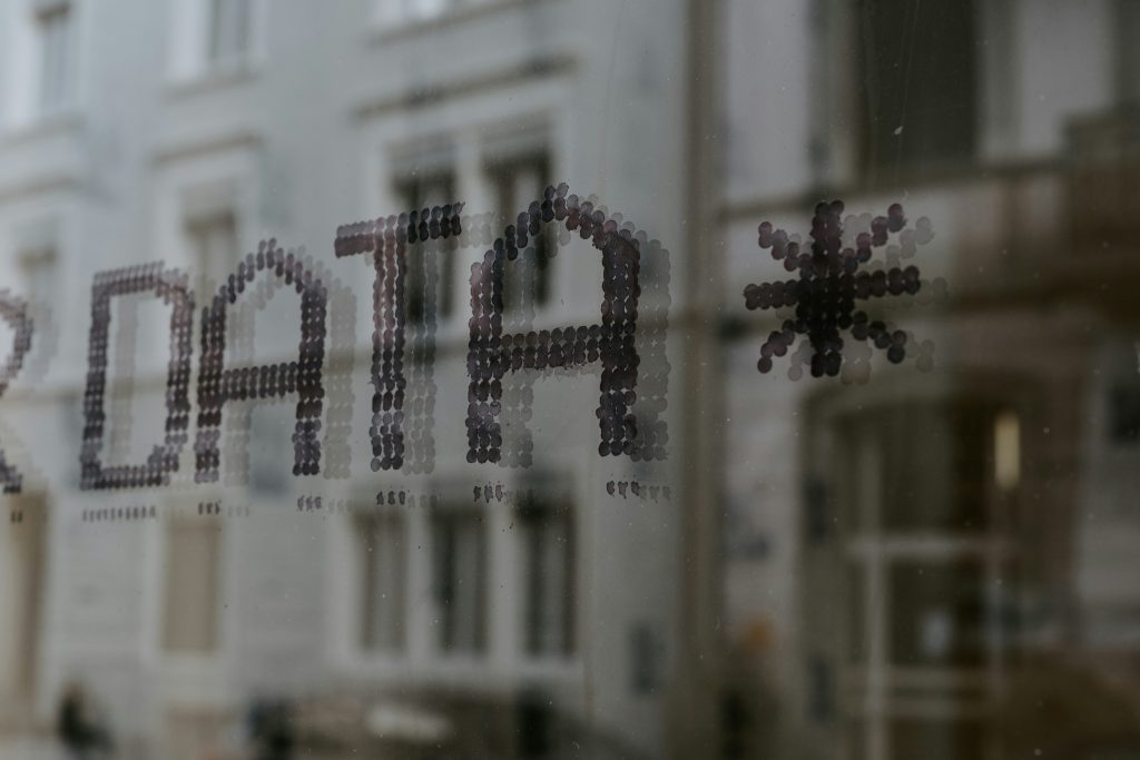 The image features the word "DATA" in large, bold, uppercase letters, followed by a symbol resembling an asterisk or snowflake. The text and symbol are composed of small, dark, circular dots, giving them a textured or pixelated appearance. They are displayed on a transparent surface, with subtle reflections visible. The background shows a blurred architectural structure with light-colored elements like windows and walls, creating a bokeh effect that emphasizes the sharp focus on the text in the foreground. The overall lighting is soft and subdued, enhancing the minimalist and modern aesthetic, making it a fitting representation of information management for small business.