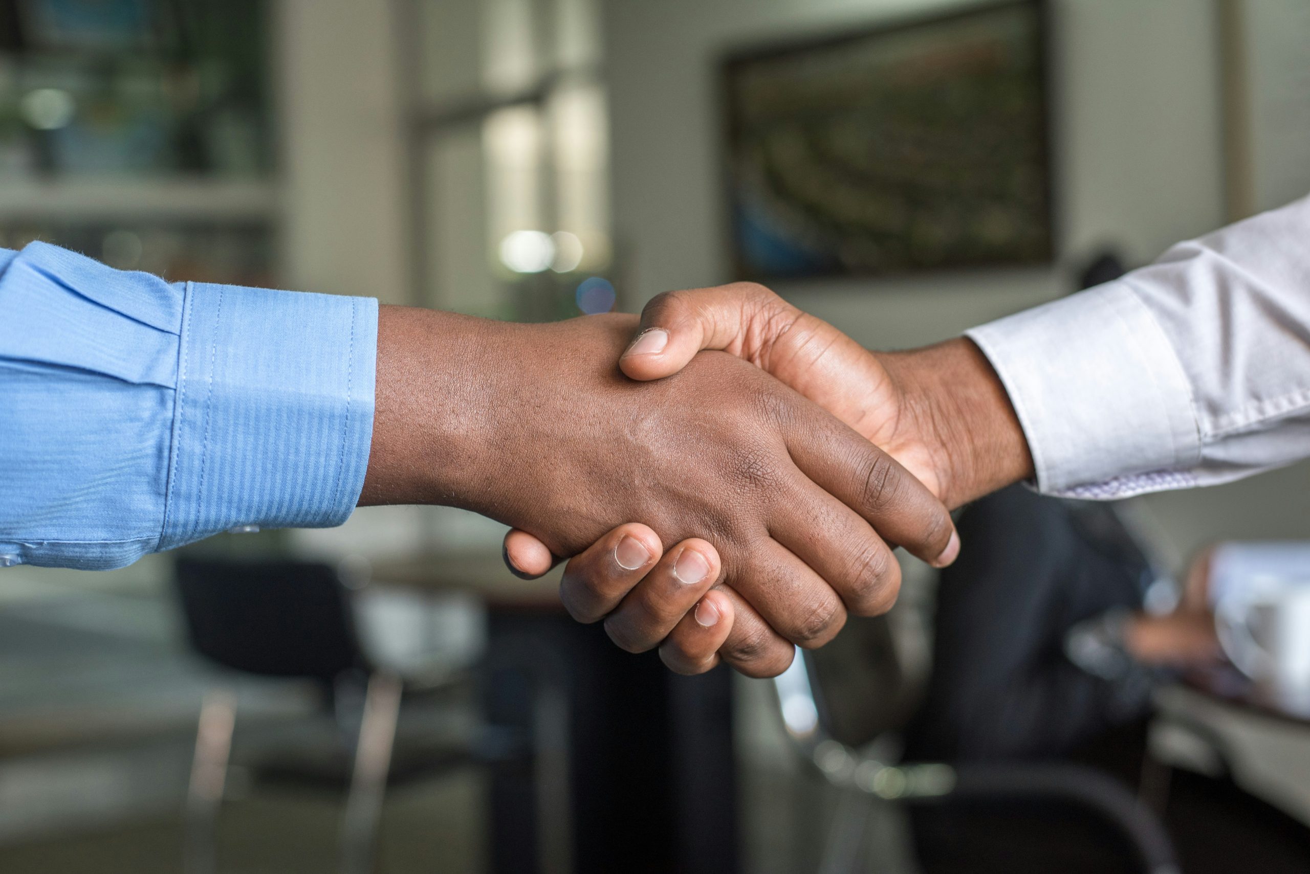 Close-up of two individuals shaking hands in a professional setting, a common sight thanks to what are angel matchmaking services. One wears a light blue striped shirt, and the other a plain white shirt. The handshake is the focal point, with a blurred office-like background featuring indistinct furniture and soft bokeh lighting.
