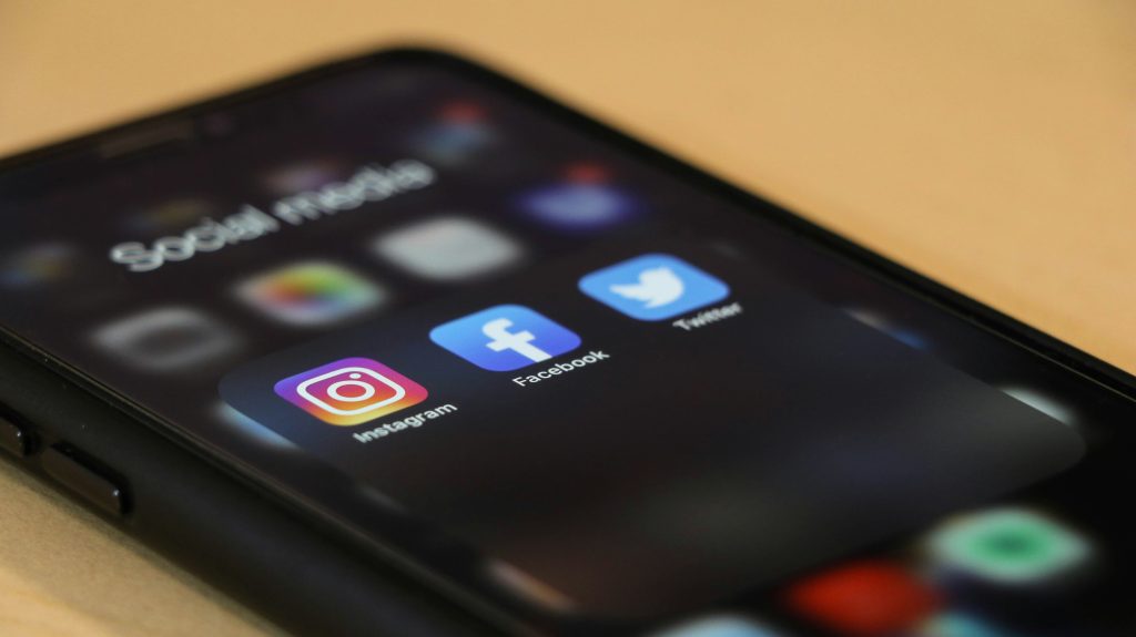 Angled close-up of a smartphone resting on a light wooden surface, displaying a home screen folder labeled 'Social media.' The screen highlights three app icons in sharp focus: Instagram (vibrant gradient square with a white camera outline), Facebook (blue square with a white 'f'), and Twitter (light blue square with a white bird). The app names are clearly visible below each icon, while the surrounding icons and background are blurred, emphasizing the social media apps and the best time to post on social media.