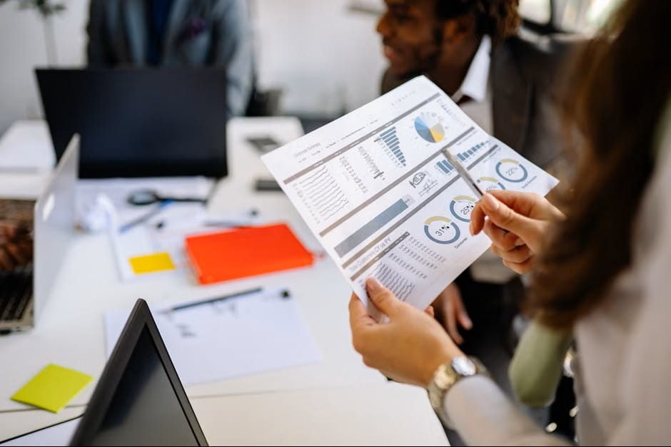 A business team reviews a printed corporate data report with pie charts and bar graphs during a collaborative office meeting.