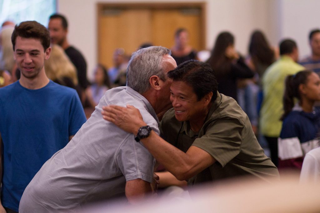Two men warmly embrace in the foreground of an indoor setting, one wearing a light gray shirt and the other a dark green shirt, both smiling. A young man in a blue shirt looks on from the left, while several other individuals are visible in the softly blurred background, engaged in conversations. The scene conveys a friendly and welcoming atmosphere, likely at a community or social gathering, such as networking events San Diego.