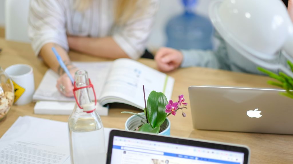 A modern workspace with a light wooden table featuring a white Apple laptop, a potted orchid with vibrant pink flowers, a glass water bottle with a red hinged cap, and a white mug with a yellow hazard symbol. A second laptop screen displays a web page, perhaps for an AI Agency, partially visible in the foreground. In the background, two individuals are blurred—one writing with a blue pen and another near a large blue water dispenser. The scene is brightly lit, blending digital tools and personal touches in a productive environment.