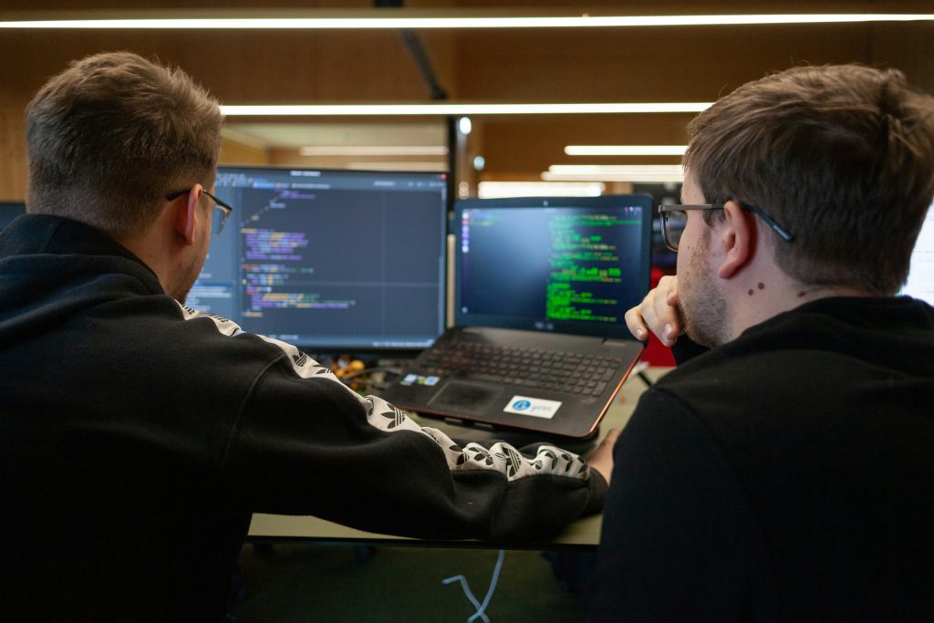 Two men collaborate in an office, focused on coding displayed on a large monitor and a laptop. The screens show programming syntax and a command-line interface, emphasizing a tech workspace and information management for small business.