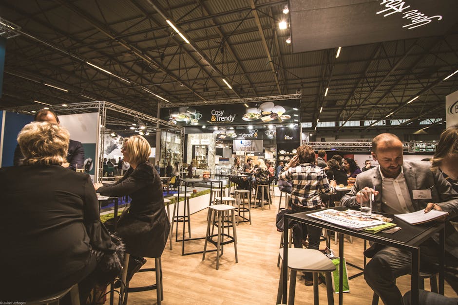 Trade show or exhibition event with attendees seated or standing around high tables. A prominent 'Cosy & Trendy' banner in the background showcases tableware items. The venue features a high industrial ceiling, warm lighting, and various exhibition booths displaying products. These free business networking events provide a great platform to connect and explore.