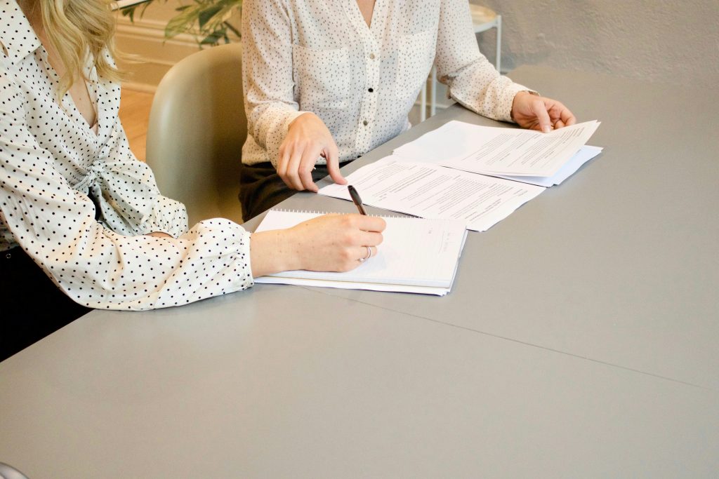 Two individuals seated at a grey table, engaged in a professional discussion. The person on the left, wearing a white polka dot shirt, is writing in a spiral-bound notebook with a pen. The person on the right, in a light-colored shirt with a subtle pattern, is reviewing a stack of documents. The table is covered with papers and a notebook, suggesting a collaborative task on topics like how to build business credit. The background is blurred, showing a light office chair and faint greenery, indicating an indoor office setting.
