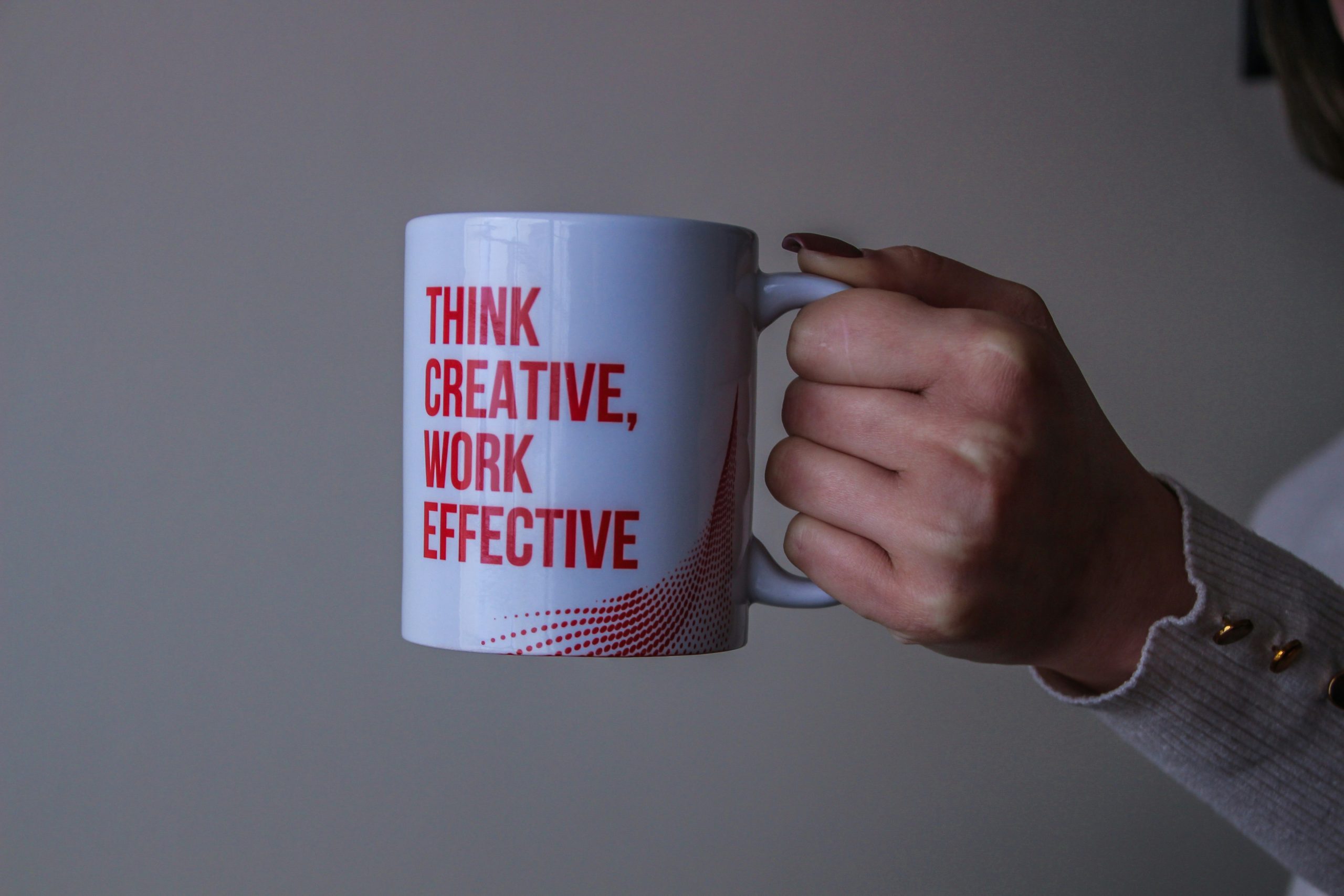 A white mug with bold red text reading 'THINK CREATIVE, WORK EFFECTIVE,' held by a hand wearing a light sweater with gold buttons, against a neutral gray background, highlighting the importance of choosing the right marketing materials photos vs videos static vs dynamic for impactful branding.