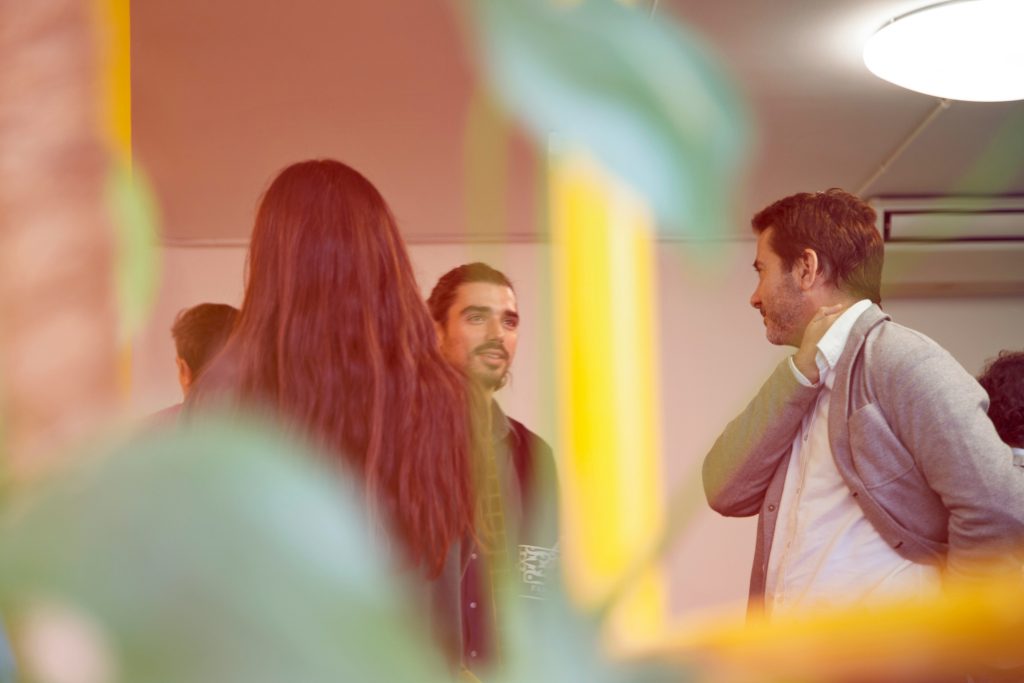 An artistic indoor scene featuring three individuals in conversation, framed by a blurred foreground of green and yellow plant-like elements. In the center, a man with dark hair tied back and a beard smiles warmly, wearing a dark shirt with a faint graphic on the sleeve. To his right, another man with short dark hair and a beard, dressed in a white collared shirt under a light gray cardigan, is seen in profile, gently touching his neck or jacket, perhaps discussing how to find angel investor Los Angeles. A third person with long reddish-brown hair is partially visible on the left, facing the group. The background includes a plain light-colored wall with a ceiling light and an air conditioning unit. Vibrant orange, red, and yellow light overlays add warmth and depth to the image, creating a candid and engaging atmosphere.
