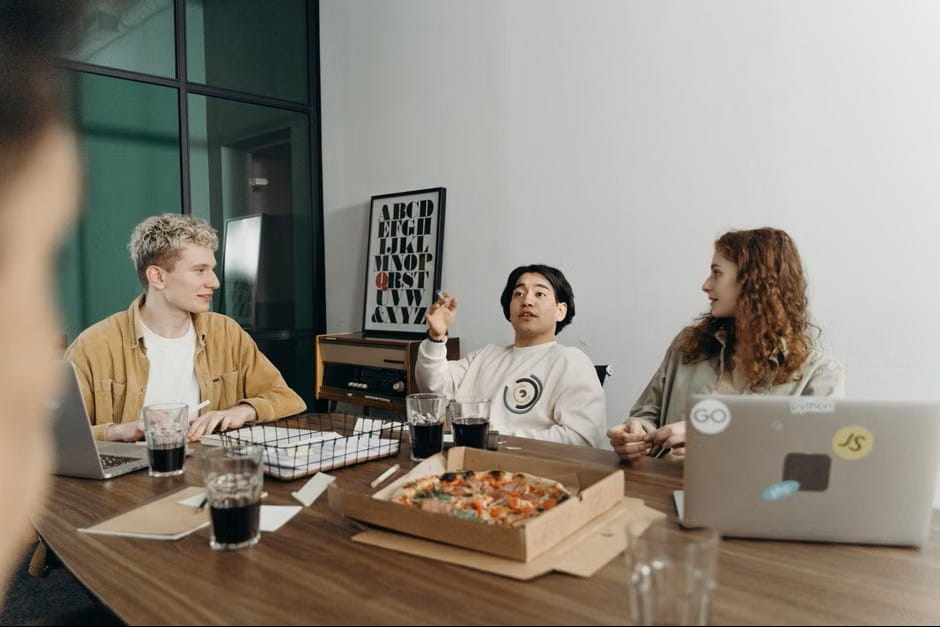 Three young tech professionals collaborate around a wooden table with laptops, pizza, and drinks, showing how to create a startup through relaxed teamwork in a modern office.